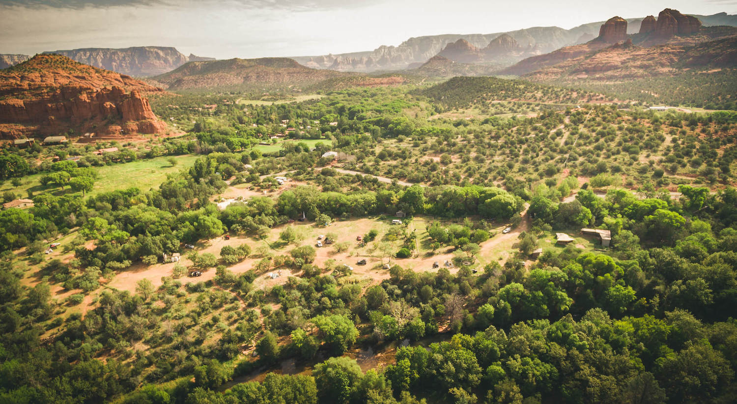 An aerial view of green fields lined with trees. In the background, red rock formations frame the horizon.